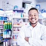 Marketing local. Na imagem, um homem farmacêutico está sorrindo de braços cruzados, em frente a uma gôndola de medicamentos.