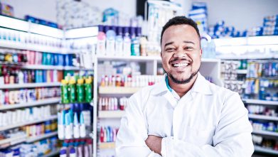 Marketing local. Na imagem, um homem farmacêutico está sorrindo de braços cruzados, em frente a uma gôndola de medicamentos.