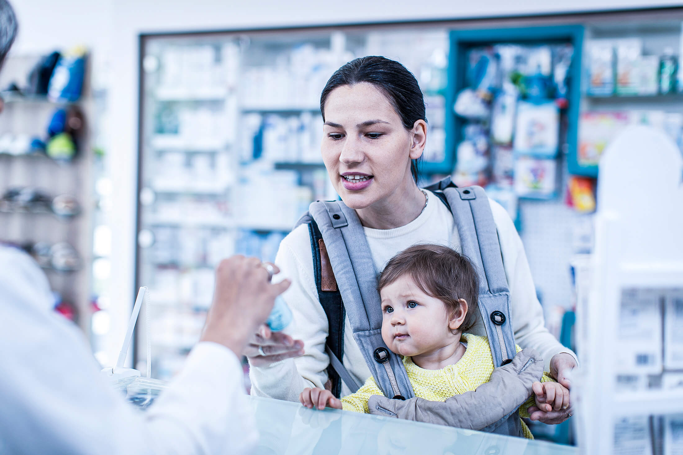 Qual o melhor horário de funcionamento da farmácia? Na imagem, uma mãe com um bebê de colo escolhe um medicamento na farmácia